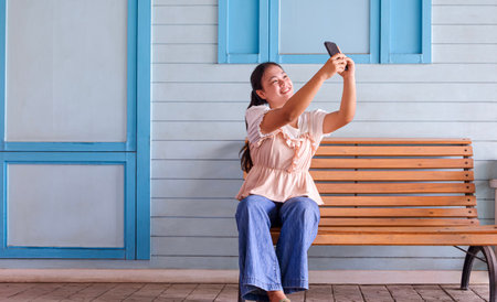 Happy Asian teenage girl is smiling and taking selfie with smartphone on wooden bench in front of pastel blue wood wall outside of vintage coffee shopの写真素材
