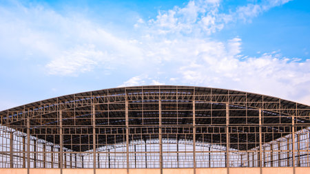 Metal structure of large warehouse industrial factory building with corrugated steel dome roof and skylights in construction site against clouds on blue sky background, Front view with copy spaceの写真素材