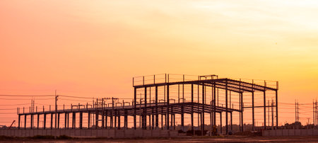 Silhouette metal structure of large warehouse factory building in construction site against orange sunset sky background, panoramic viewの写真素材