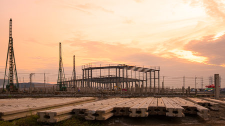 Silhouette of industrial warehouse factory building with pile driver machines and many precast concrete piles on the ground in construction area against orange sunset sky background in the eveningの写真素材