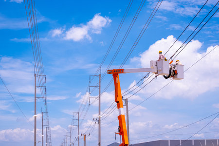 Two electricians on bucket boom truck are connecting electrical cable lines in industrial settlement area against blue sky background, low angle view with copy spaceの写真素材