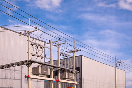 Two factory industrial production buildings with overhead cable tray and electric pole with power lines against blue sky background, low angle and perspective side viewの写真素材
