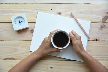 Woman's hand with a white cup of coffee and notebook with wooden background. The concept of a coffee breakの写真素材