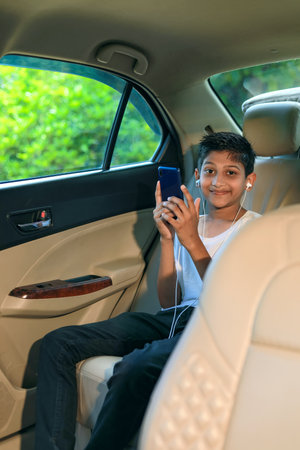Cute indian child sitting in car and using smart phone and headphones gadget