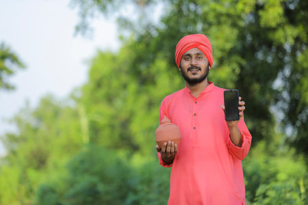 Young indian farmer holding clay piggy bank and showing smart phone in handの写真素材