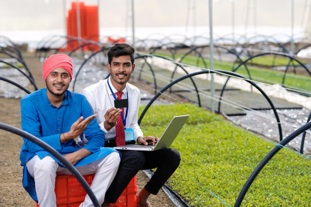 Young indian agronomist showing some information to farmer in laptop at greenhouse