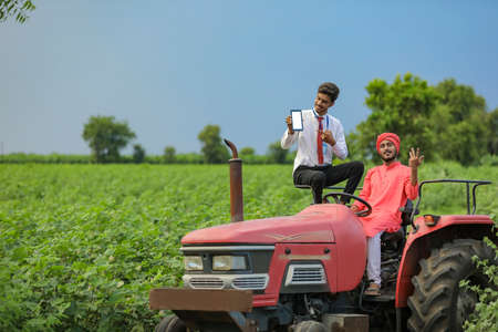 Young indian farmer and bank officer sitting on new tractor and officer showing mobile screen at agriculture fieldの写真素材