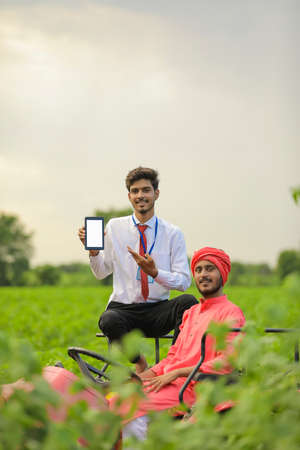 Young indian farmer and bank officer sitting on new tractor and officer showing mobile screen at agriculture fieldの写真素材