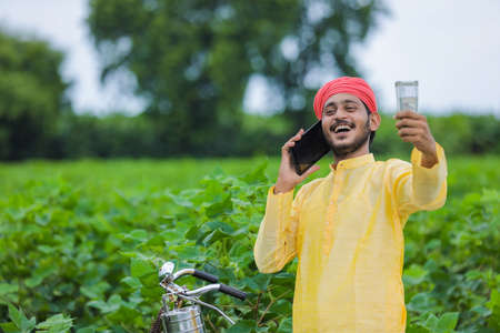 Young indian farmer talking on mobile phone and showing money at cotton field