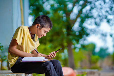 Indian school boy holding phone distance learning class using mobile application, watching online lesson, video calling in app making notes studying at homeの写真素材