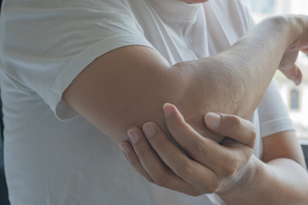 An Asian man holds his right elbow with visible tension, hinting at localized pain or strain. The...