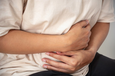 The person grips their abdomen, suggesting abdominal pain that may result from gastrointestinal distress, bloating, or inflammation. The indoor setting implies a home or clinic.の写真素材