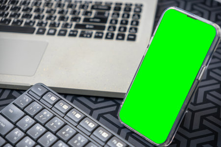 Modern tech workspace with laptop, green-screen smartphone, and backlit keyboard on a patterned desk. Ideal for tech project ads, device integration, digital innovation. Copy spaceの写真素材