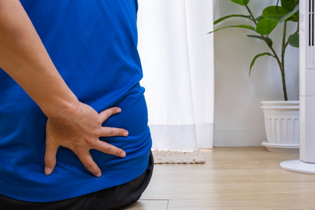Senior man holding lower back in pain while standing indoors. Calm setting with copy space for...
