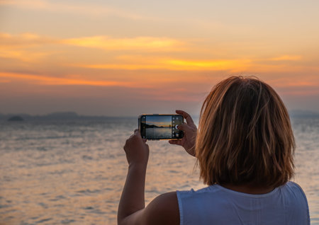 Asian woman capturing sunset scenery with smartphoneâ€”ideal for promoting scenic travel,...