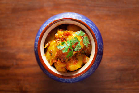 Close-up of Indian vegetarian dish of spicy Potato and Tomato curry garnished with green coriander fresh leaves. Served in ceramic handi over wooden background.の写真素材
