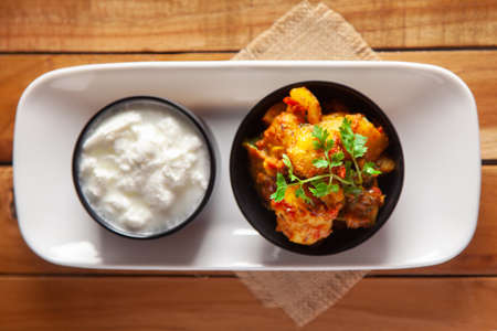 Close-up of Indian vegetarian dish of spicy Potato and Tomato curry garnished with green coriander fresh leaves with curd, over wooden background.の写真素材