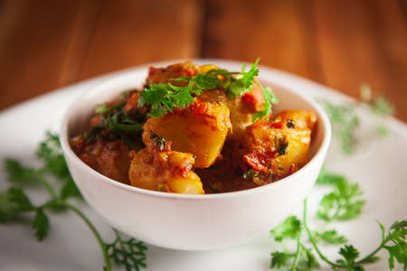 Close-up of Indian vegetarian dish of spicy Potato and Tomato curry garnished with green coriander fresh leaves. Served in a white ceramic bowl over wooden background.の写真素材