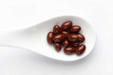 Macro Close up of medicinal or herbal dark red-brown pill on a white ceramic spoon. Top view, white background.の写真素材