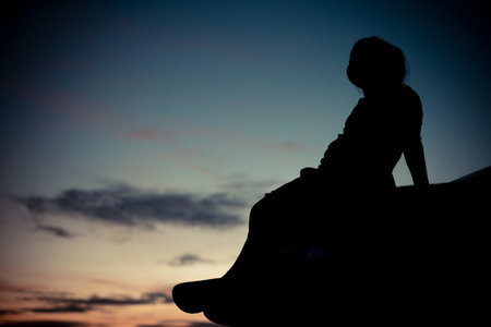 Silhouette of a little girl sitting on the edge of a cliff at sunsetの写真素材
