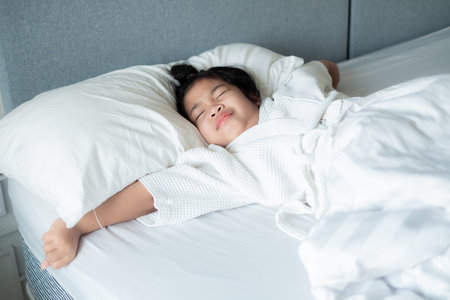 young asian girl stretches and smiles in bed after waking up, wearing a white bathrobe and enjoying a peaceful, cozy morning in a bright and comfortable bedroomの写真素材