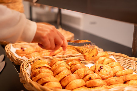 A person using tongs to pick a freshly baked pastry from a basket at a bakery or breakfast buffet, highlighting golden brown texture and warm, inviting atmosphere.の写真素材