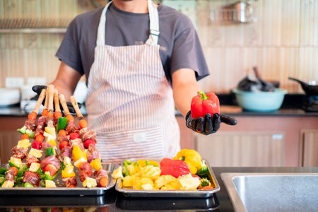 Chef holding a red bell pepper while preparing colorful meat and vegetable skewers, showcasing fresh ingredients and vibrant food preparation in a home kitchen setting.の写真素材