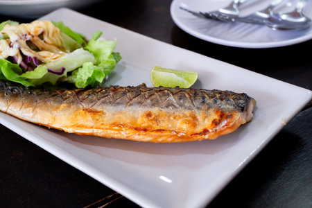 Grilled mackerel served on a white plate with fresh green salad, topped with creamy dressing and a...