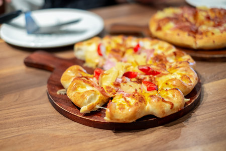 Hot baked cheese pizza with ham, pineapple, and bell pepper served on a wooden tray in a restaurant. Freshly cooked delicious pizza ideal for food, lunch, dinner, or Italian cuisine concepts.の写真素材