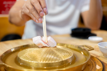Close-up of a hand using chopsticks to place thin slices of raw pork or beef meat onto a hot brass Mookata or Asian BBQ grill plate. Asian dining and cooking experience at a restaurant dinnerの写真素材