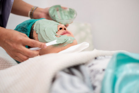 Asian woman receiving professional facial treatment at beauty spa salon. Beautician applying green natural clay mask on face with spatula. Wellness, skincare and rejuvenation conceptの写真素材