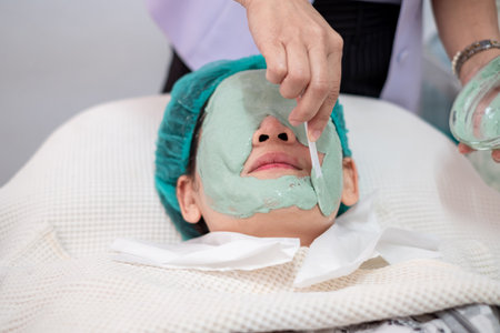 Asian woman receiving professional facial treatment at beauty spa salon. Beautician applying green natural clay mask on face with spatula. Wellness, skincare and rejuvenation conceptの写真素材