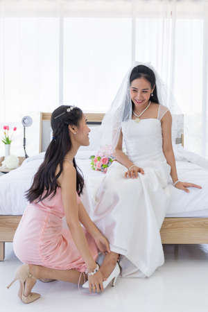 An Asian bride in a white wedding dress is sitting on the bed and a bride's friend is helping her to wear high heels.の写真素材