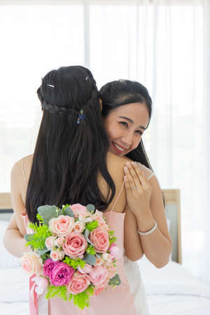 An Asian bride in a white wedding dress embraces her friend in a pink dress with a smile, then shows a diamond ring on her left finger and on the other hand, holding a beautiful bouquet of flowers.の写真素材