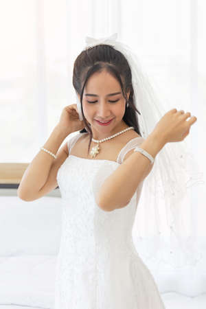 An Asian bride dressed in a white wedding dress stands in a cute and bright smile beside the bed in the room.の写真素材