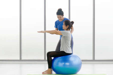 A female trainer is teaching a middle-aged Asian female customer to do yoga sitting on a rubber ball in a gymnasium.の写真素材