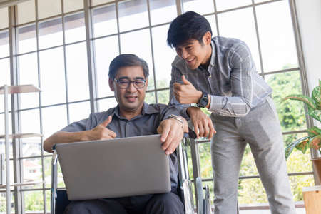 A middle-aged Asian father sits in a wheelchair and has a son next to him chatting on video calls on a laptop.の写真素材