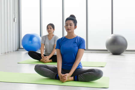 Two middle-aged Asian women doing yoga sitting on a rubber mat in a gym.の写真素材
