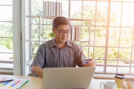 A middle-aged Asian man is using his credit card to shop online via a laptop on his desk.の写真素材