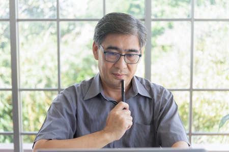 An elderly Asian man making a thinking gesture in his hand holding a penの写真素材