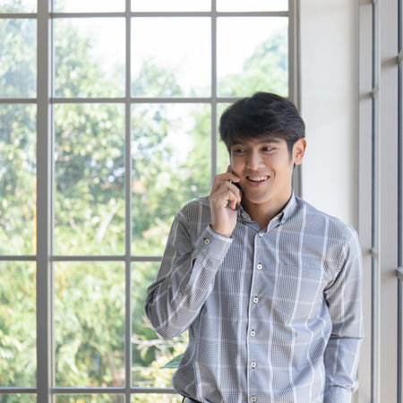 A young Asian businessman stands on the balcony of a window talking on the phone.の写真素材