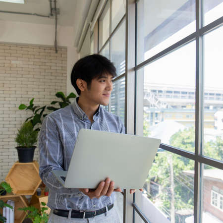 A young Asian businessman with a laptop standing by a glass window