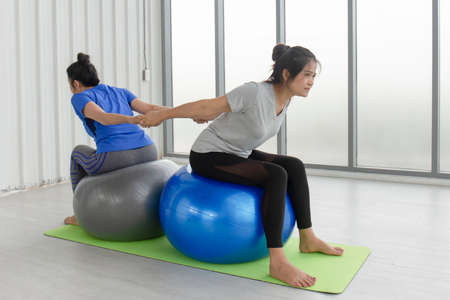 Two middle-aged Asian women doing yoga sitting on a rubber ball in the gym.の写真素材