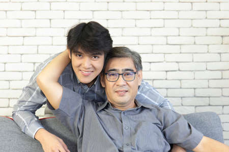 A middle-aged father and an Asian son hugged each other on the sofa with a happy smile.の写真素材