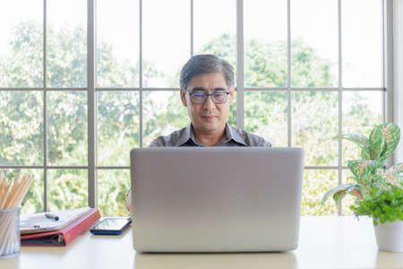 An elderly Asian man is learning how to trade online with a laptop on his desk.の写真素材