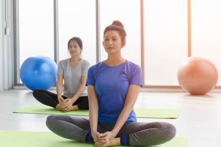 Two middle-aged Asian women doing yoga sitting on a rubber mat in a gym.の写真素材