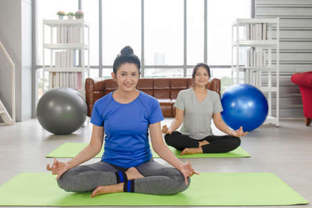 Two middle-aged Asian women are doing yoga on a rubber mat in her home.の写真素材