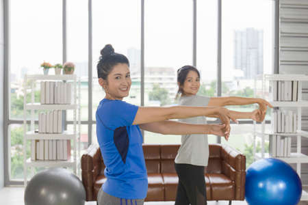 Two middle-aged Asian women are doing yoga inside their home.