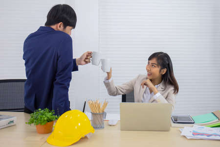 A young, professional young Asian businesswoman is sitting at her desk and bumping a cup of coffee with a male office worker working together.の写真素材