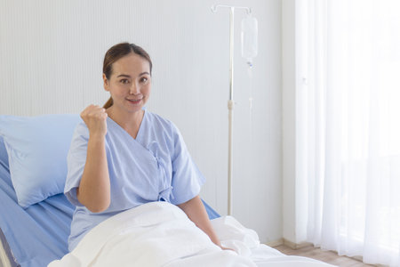 Asian female patients lie on hospital beds, raise hands, fists, show confident symptomsの写真素材